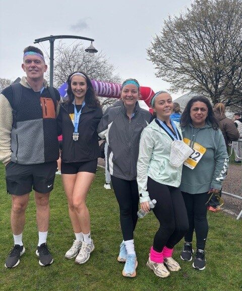 Close-up group photo of event participants wearing race bibs and blue headbands, standing among a crowd of runners outdoors, with trees and spectators visible behind them.